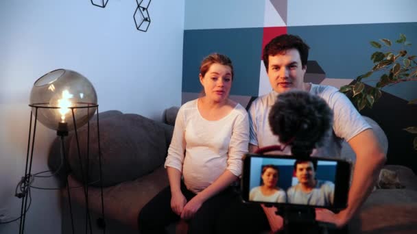 Casal jovem esperando um bebê é entrevistado na frente da câmera do smartphone em pé no suporte - Filmagem, Vídeo