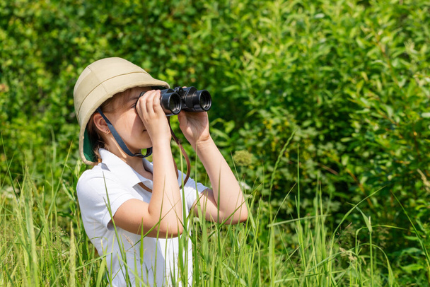 Preteen girl wearing cork helmet standing in grass looking through binoculars. Discovery concept - Foto, immagini
