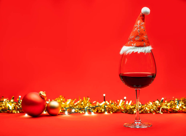 Glass of red wine with christmas baubles tinsel and lights on red background - christmas drink concept - Photo, Image