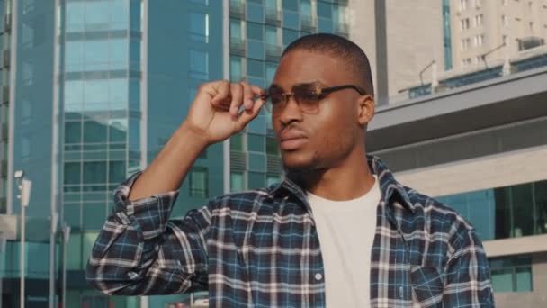 Muotokuva mietteliäs järkyttynyt afrikkalainen amerikkalainen mies aurinkolasit nuori tyylikäs kaveri seisoo kaupungin taustalla rakennuksen ajattelu katselee ympärilleen huolestunut ilme kasvot, mies turisti menetetty kadulla yksin - Materiaali, video