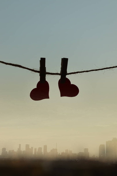 Dos corazones rojos colgando de una cuerda atada con alfileres de clavijas de ropa sobre el amanecer naranja del cielo matutino y la vista borrosa del edificio de rascacielos de la ciudad. Momento de amor romántico, feliz día de San Valentín concepto de celebración - Foto, imagen
