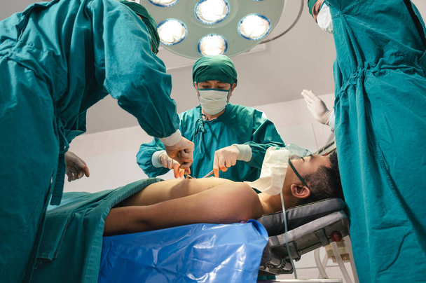 Ázsiai sebészek sebészeti köpenyben sebészeti beavatkozást végeznek egy súlyosan sérült betegen a műtőben a kórházban. - Fotó, kép