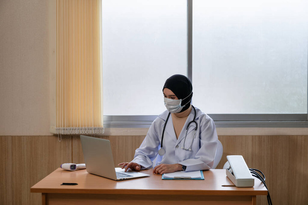 Doctora musulmana usando hijab en uniforme médico usando portátil y esfigmomanómetro, termómetro infrarrojo en el escritorio en el hospital - Foto, Imagen