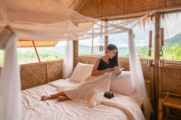Piękna młoda Azjatka odpoczywa i czyta książkę na łóżku w krytym strzechą domku pośród gór na wakacjach - Zdjęcie, obraz