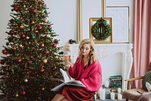 Schöne blonde Frau in einem gemütlichen weinroten Pullover mit einem Glas Champagnerrose feiert das neue Jahr in der Nähe des Weihnachtsbaums, in einem geschmückten Interieur.. - Foto, Bild