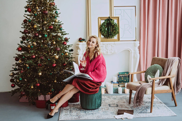 Mulher loira bonita em uma camisola borgonha aconchegante com um copo de champanhe rosa celebra o novo ano perto da árvore de Natal, em um interior decorado.Foco seletivo suave. - Foto, Imagem