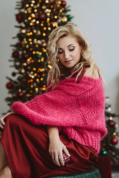 Mooie blonde vrouw in een gezellige bordeaux trui en een zijden jurk viert het nieuwe jaar tegen de achtergrond van de bokeh lichten van een kerstboom.Zachte selectieve focus. - Foto, afbeelding