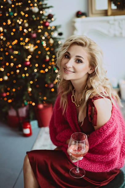 Schöne blonde Frau in einem gemütlichen weinroten Pullover mit einem Glas Champagnerrose feiert das neue Jahr in der Nähe des Weihnachtsbaums, in einem geschmückten Interieur.. - Foto, Bild