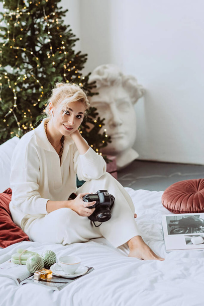 Jonge mooie vrouw in een modern nieuwjaarsinterieur, in een witte slaapkamer, tegen de achtergrond van bokeh lichten van een kerstboom met een camera in haar handen. Zachte selectieve focus. - Foto, afbeelding