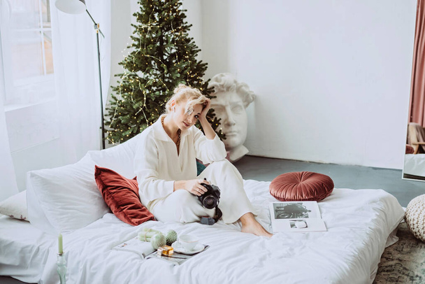 Young beautiful woman in a modern New Year's interior, in a white bedroom, against the background of bokeh lights of a Christmas tree with a camera in her hands. Soft selective focus. - Fotó, kép