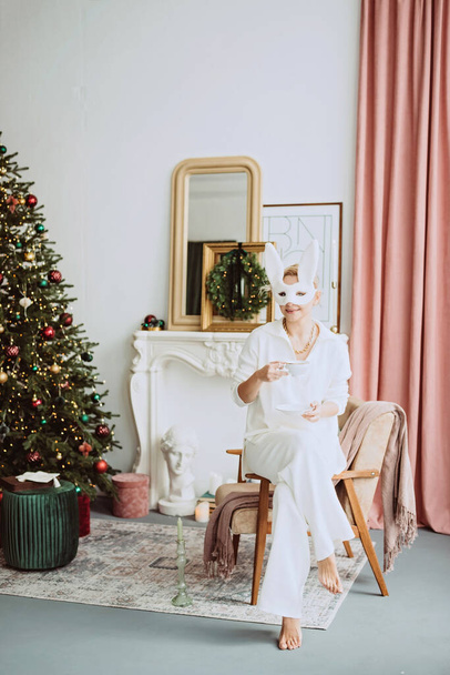 カーニバルのウサギのマスクと白い衣装を着た美しい若い女性は、現代のインテリアでクリスマスツリーの近くに新年を祝います。インスピレーション。柔らかい選択的フォーカス. - 写真・画像