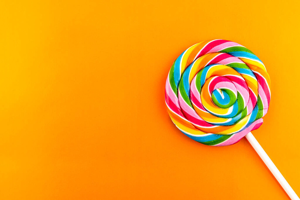 Sweet colorful spiral shaped lollipop lying in lower right corner, isolated on orange background, top view. - Photo, image