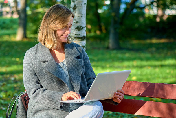 Una mujer con una computadora portátil está sentada en un banco del parque en un día soleado de verano - Foto, Imagen