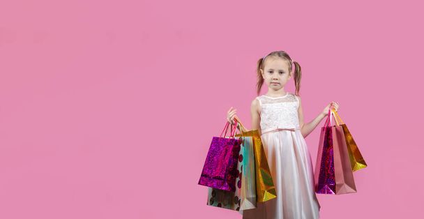 Χαριτωμένο μικρό καυκάσιο κορίτσι σε ένα κόμμα φόρεμα σε ροζ φόντο ευτυχής απολαμβάνοντας ψώνια κρατώντας πολύχρωμες τσάντες - Φωτογραφία, εικόνα