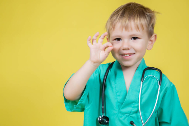 Portrait of cute little caucasian boy dressed in doctors green showing okay ok gesture on yellow background - Foto, Bild