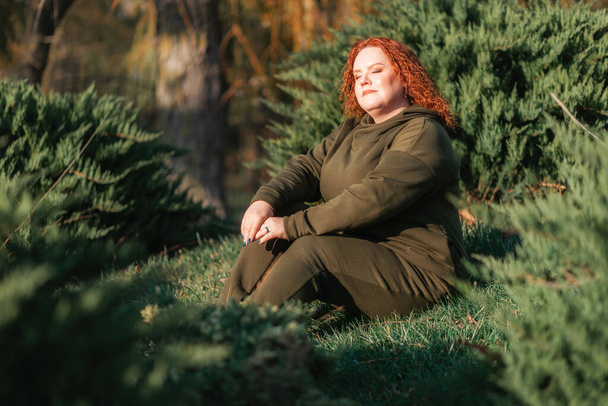 Eine übergewichtige schöne Frau wärmt sich in der Sonne, sitzt auf dem Rasen im Park. Das Konzept von Psychologie und Frieden. - Foto, Bild