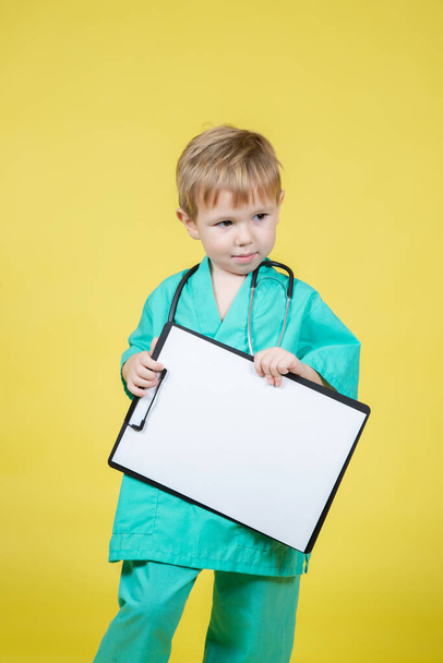 Portrait of little caucasian kid dressed in doctors green coat holds tablet in hand isolated on yellow - Фото, зображення