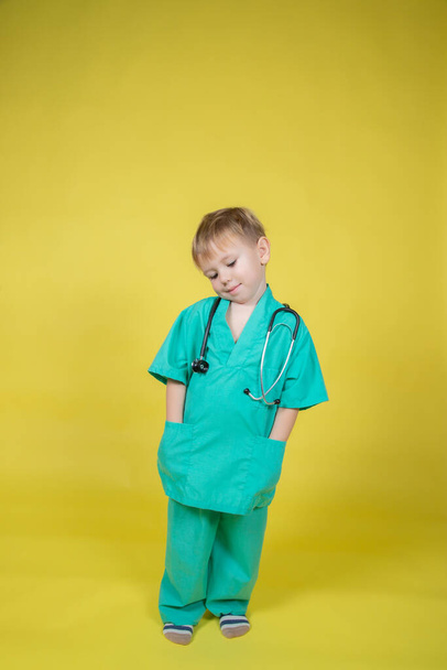 Portrait of little caucasian boy dressed in doctors green coat posing isolated on yellow - Fotoğraf, Görsel