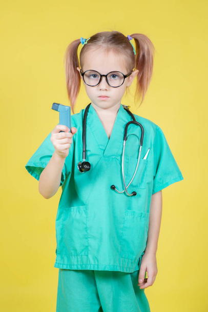Portrait of little caucasian girl dressed in doctors green coat with an asthma inhaler in hand on yellow background - Φωτογραφία, εικόνα