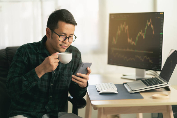 Asian man trader drinking coffee and sitting at home office in front of monitors with cryptocurrency graph holding smartphone browsing application monitoring cryptocurrency price - Photo, Image