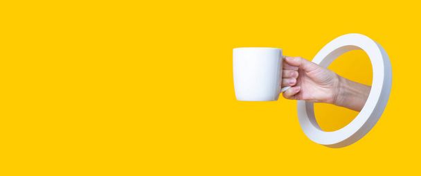 Mão feminina segurando uma xícara branca em um buraco redondo em um fundo amarelo. Banner - Foto, Imagem