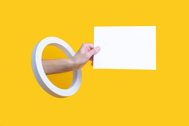 Mano femenina sosteniendo una hoja en blanco en un agujero redondo sobre un fondo amarillo - Foto, imagen