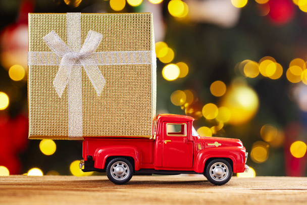 Красный ретро-игрушечный грузовик несет подарок на фоне рождественской елки с рождественскими огнями. Концепция доставки товара и поздравительная открытка с Новым годом. - Фото, изображение
