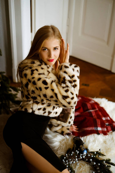 portrait of a young caucasian woman with long blond hair wearing a leopard fur coat, a beautiful slim girl in a dress and fur coat is sitting on the floor near the Christmas tree, bright lady with - Photo, image