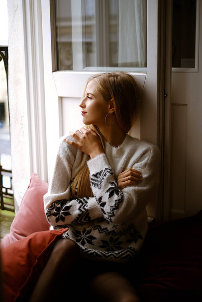A young caucasian woman in a warm sweater sits at an open window, a beautiful lady is wrapped up in a patterned sweater in a stylish apartment, a blonde in black tights and a light top sits on the - Photo, Image