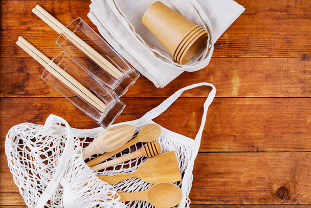 Wooden cutlery kitchenware and mesh bag on wooden table. Bamboo straws in empty glasses on a wooden boards. Disposable paper cups and linen shopping bag. Zero waste concept - Foto, immagini
