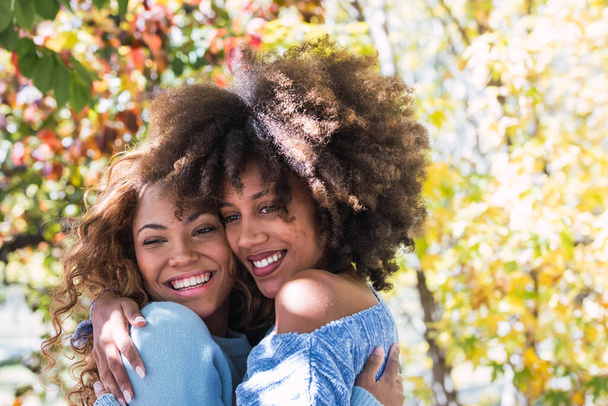 Primer plano de dos amigas afro americanas felices abrazándose y riéndose en el parque. Alegre afro negro amigos con el pelo rizado sonriendo con amor y dando cálido abrazo - Foto, imagen