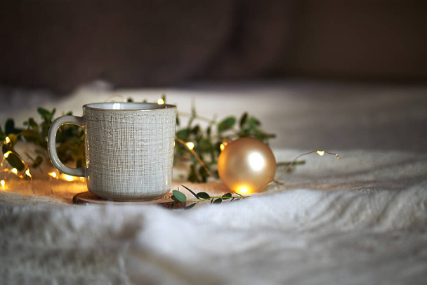 Κεραμική κούπα καφέ σε ένα μπεζ πλεκτό φόντο με φύλλα ευκαλύπτου και χριστουγεννιάτικα φώτα γιρλάντα, έννοια Χριστούγεννα ζεστή χειμωνιάτικη μέρα στο σπίτι hugge, selecive εστίαση, αντίγραφο χώρου - Φωτογραφία, εικόνα