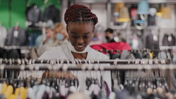 Fiatal elegáns afro-amerikai lány vásárló a bevásárló központban ruhaüzlet választ ruhát keres gyönyörű ruhákat talál kabát fut át állvány mosolygós elégedett megállapítás - Felvétel, videó