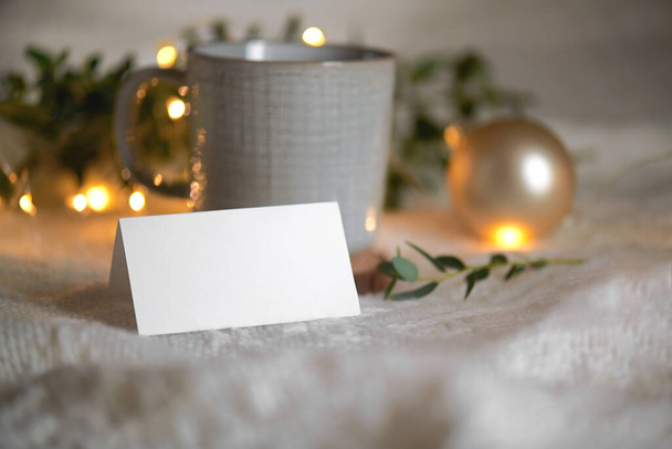 Christmas empty stationery card mockup with eucaliptus leaves, mug and light garland on knitted beige cozy background, design element for invitation, thank you or greeting card, business card, rsvp - Photo, Image