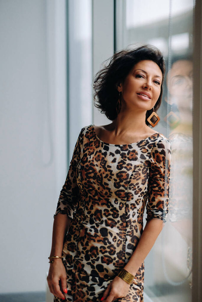 a brunette woman in a tiger dress stands near a large window. - Fotografie, Obrázek