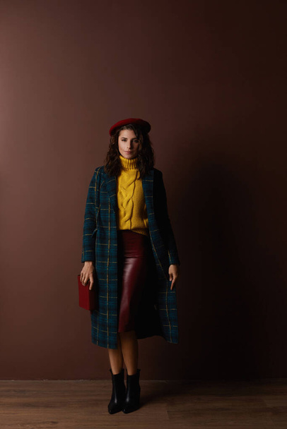 Mid-aged women wearing a checkered coat, yellow sweater, heels, midi skirt, and beret posing indoors against a beige wall - Фото, изображение
