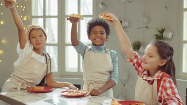 Ομαδικό πορτρέτο μικρών πολυεθνικών παιδιών σε ποδιές κρατώντας φέτες φρεσκοψημένης πίτσας και χαμογελώντας στην κάμερα ενώ ποζάρουν στην κουζίνα κατά τη διάρκεια του μαθήματος μαγειρικής - Πλάνα, βίντεο