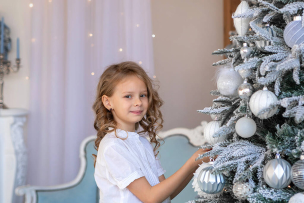 A blonde girl with curly hair decorates a Christmas tree - Foto, afbeelding
