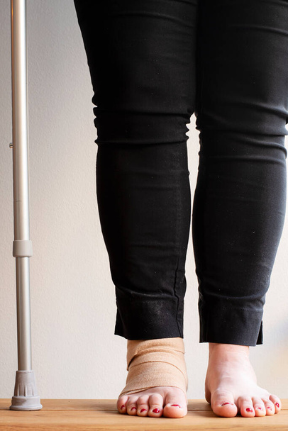 Legs of a person with one foot wrapped with an elastic bandage. Next to the person is a crutch, a mobility aid to be used as support due to injury or disability. - Photo, Image