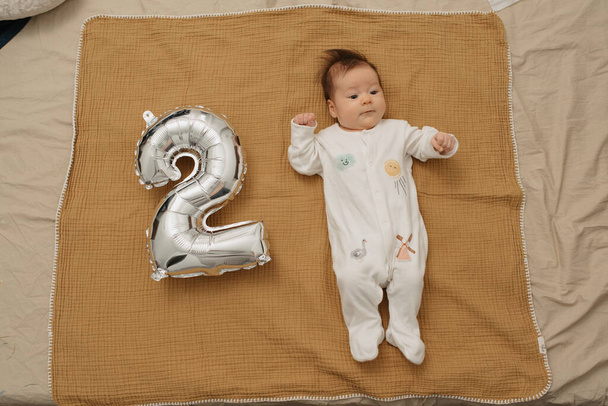 Un bebé serio está acostado en una manta de muselina cerca de un globo de lámina de plata en forma de 2. Una niña vestida de una sola pieza celebra sus dos meses. - Foto, Imagen