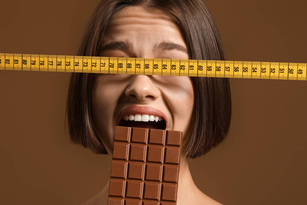 Stresowana młoda kobieta ze słodką czekoladą i taśmą pomiarową na tle koloru. Koncepcja diety - Zdjęcie, obraz