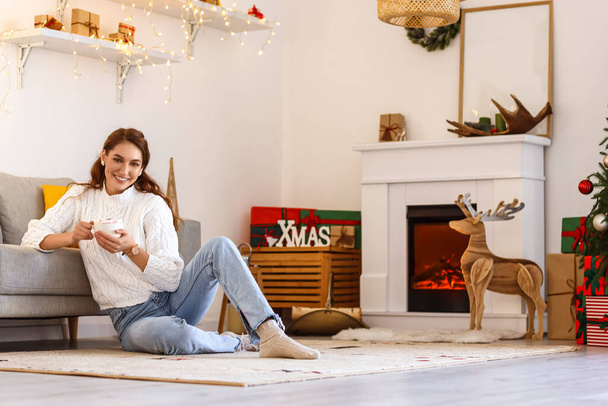Красивая молодая женщина с чашкой горячего шоколада дома в канун Рождества - Фото, изображение