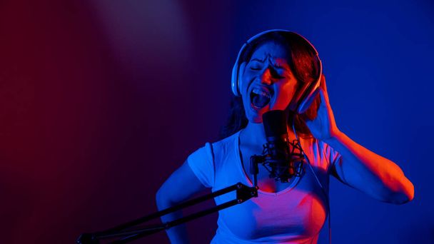 Caucasian woman in glasses and headphones sings into a microphone in neon light on a black background. An emotional girl is recording a song in a recording studio - Фото, изображение