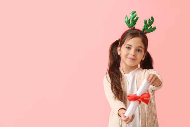 Grappig klein meisje met rendieren hoorns en scroll op kleur achtergrond - Foto, afbeelding