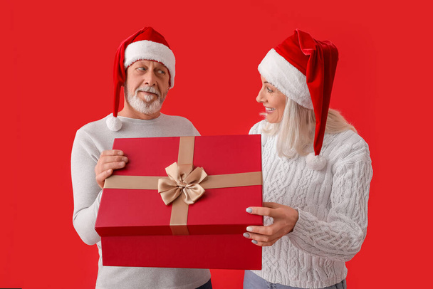 Ευτυχισμένο ώριμο ζευγάρι σε Σάντα καπέλα και με χριστουγεννιάτικο δώρο στο φόντο χρώμα - Φωτογραφία, εικόνα