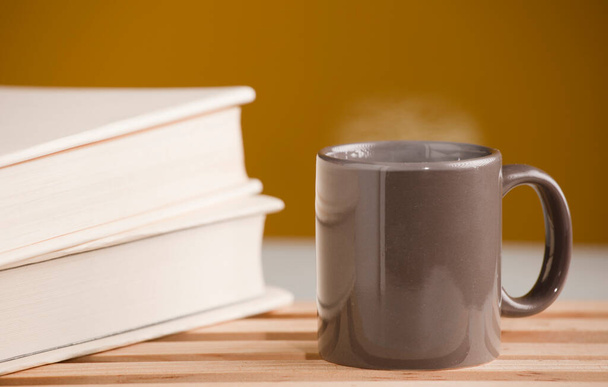 Braune Tasse Kaffee oder Tee mit heißem Dampf, auf dem Tisch, mit Buch und braunem Hintergrund. - Foto, Bild