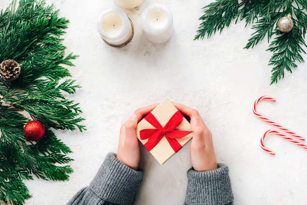 Lapsen käsissä on minimalistinen joululahjapakkaus, joka on koristeltu punaisella nauhalla. Fir oksat, kynttilät ja karkkia keppejä valkoisella kuvioitu tausta. Top view, tasainen lay. - Valokuva, kuva