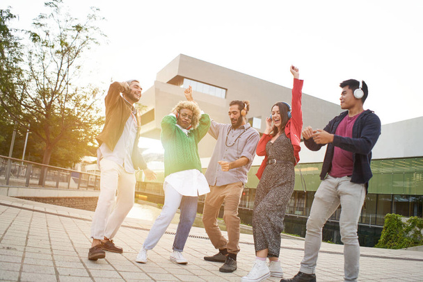 Група колег святкує. Веселі люди танцюють на свіжому повітрі під час заходу сонця. Молоді тисячоліття з навушниками слухати музику
. - Фото, зображення