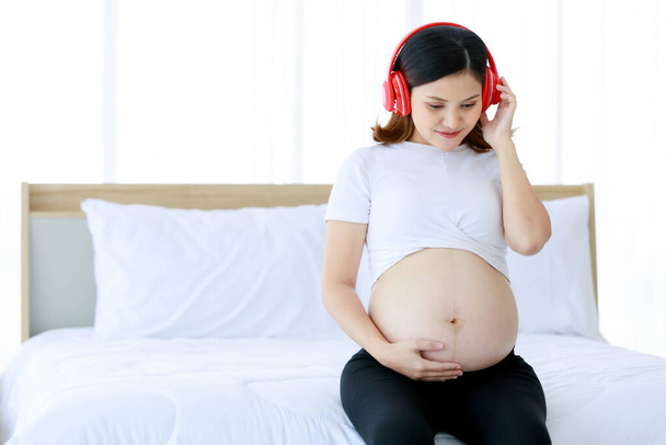 Ázsiai fiatal boldog gyönyörű terhesség nő anya visel nagy piros fejhallgató hallgat lágy dal zene ül pihentető egyedül az ágyban a hálószobában gazdaság átölelő meztelen has has. - Fotó, kép