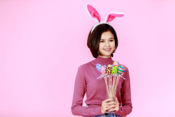 Studioaufnahme von asiatischen jungen glücklichen ziemlich kurzen Haaren Teenager Mädchen Modell mit Hasenohren Stirnband stehend lächelnd mit traditionellen Festival bunt bemalten Ostereiern auf Stöcken auf rosa Hintergrund. - Foto, Bild